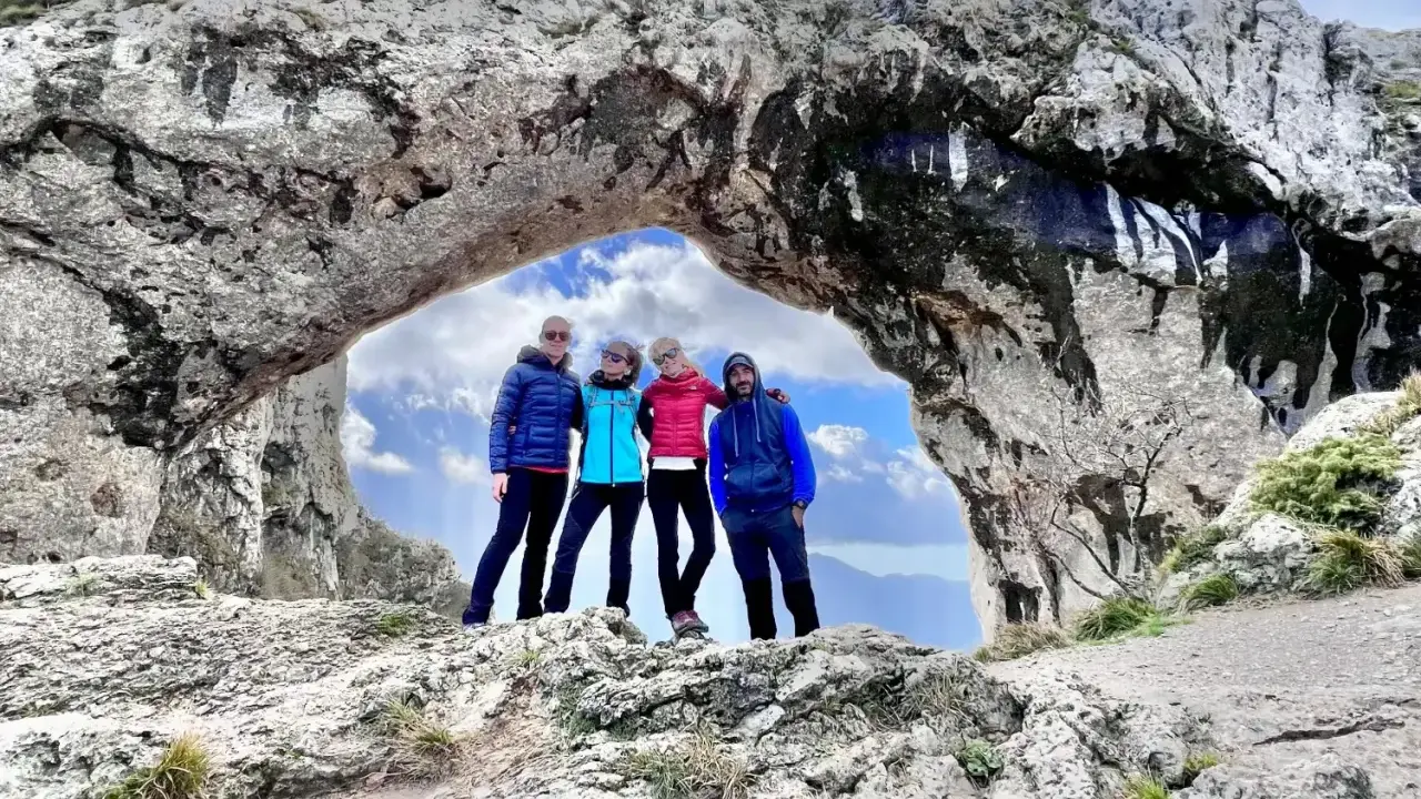 Quattro amici si godono una giornata di camminare in montagna, posando sotto un arco naturale di roccia con un cielo azzurro e nuvoloso.