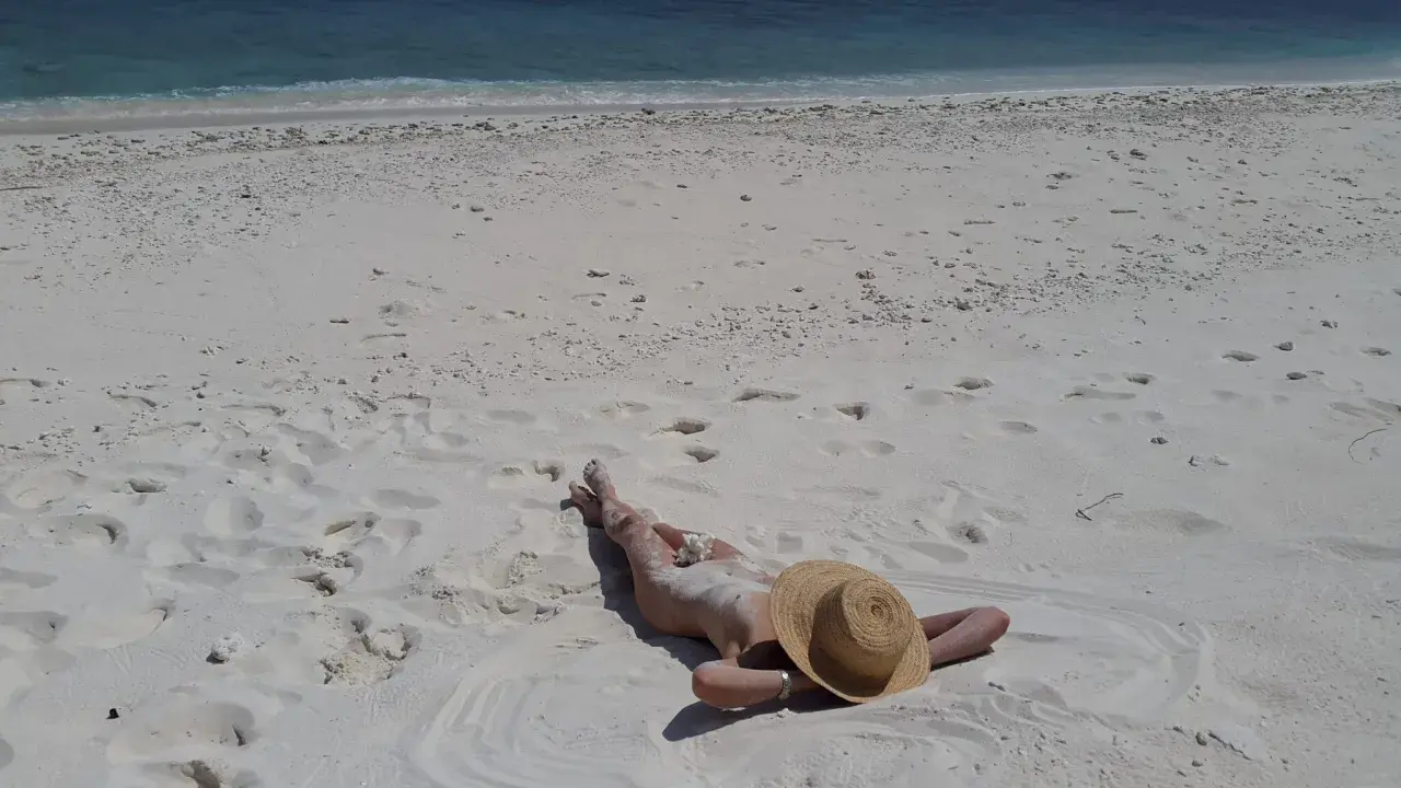 Persona distesa su sabbia bianca, con cappello di paglia, vicino al mare turchese. Un'immagine che evoca la pace delle spiagge naturiste in Italia.