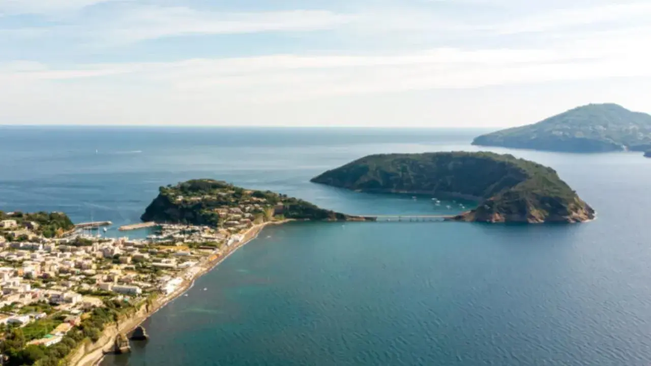 Panoramica di un'isoletta tra Napoli e Ischia, con un borgo costiero, un porto turistico e un ponte che collega a un'altra isola verdeggiante.