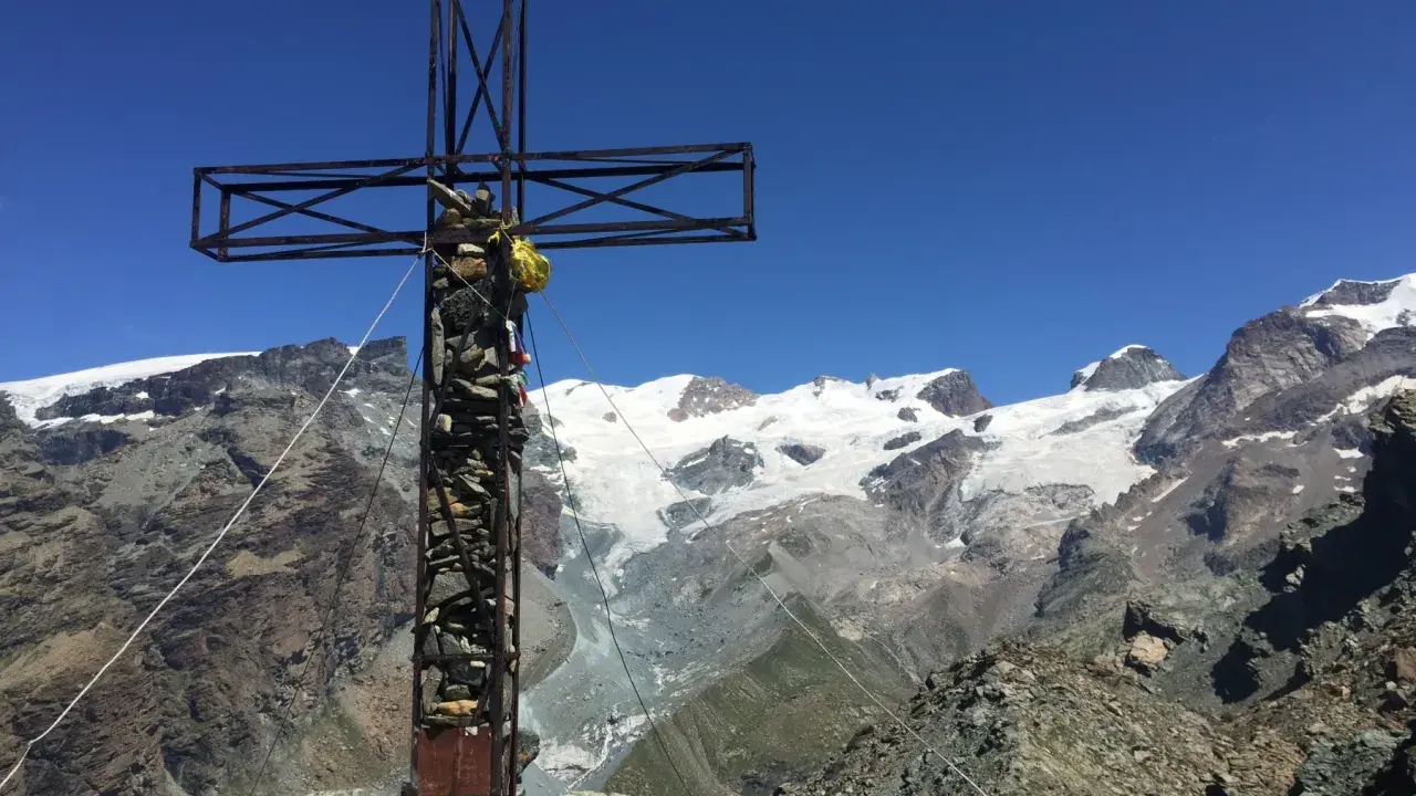 Croce metallica e di pietra in cima a una montagna, con un ghiacciaio e vette innevate sullo sfondo. Il **palon di Resy** domina il panorama.