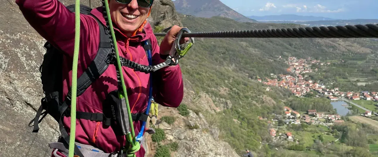 Donna sorridente si aggrappa a un cavo d'acciaio, godendosi un'avventura sulle ferrate Val di Susa con un panorama mozzafiato.