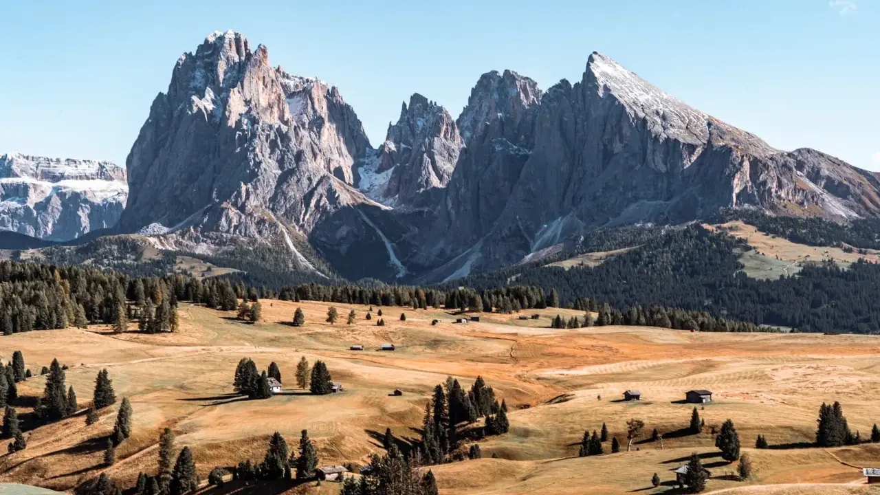 Un paesaggio mozzafiato delle Dolomiti più belle, con cime imponenti, prati dorati e boschi verdi. Piccole baite sparse creano un'atmosfera idilliaca.