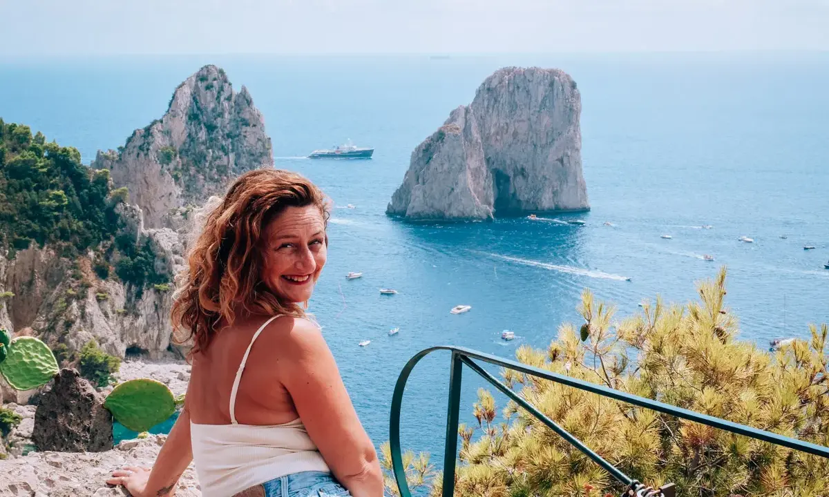Donna sorridente con vista sui Faraglioni, un'ispirazione per come visitare Capri. Mare azzurro e barche in lontananza.