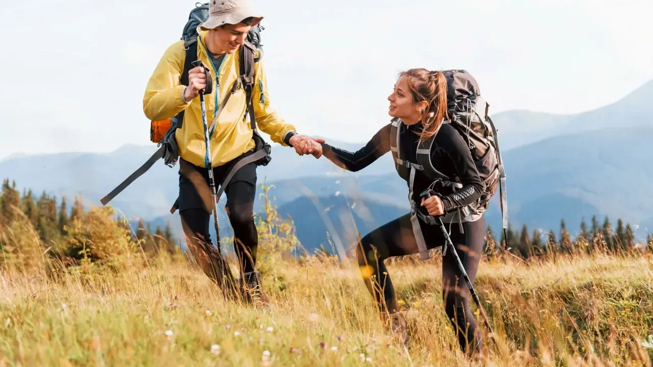 Coppia di escursionisti si aiutano a vicenda su un sentiero di montagna, con zaini e bastoncini da trekking.