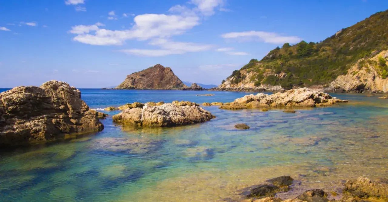 Acque cristalline e rocce scolpite dal mare a Porto Ercole, con montagne verdi sullo sfondo e un cielo azzurro punteggiato di nuvole.