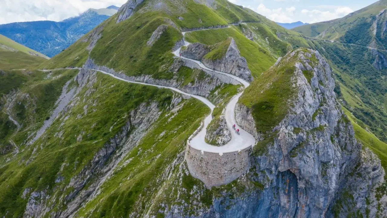Un tortuoso cammino del sale si snoda tra montagne verdi, con ciclisti che affrontano tornanti mozzafiato su un sentiero panoramico.