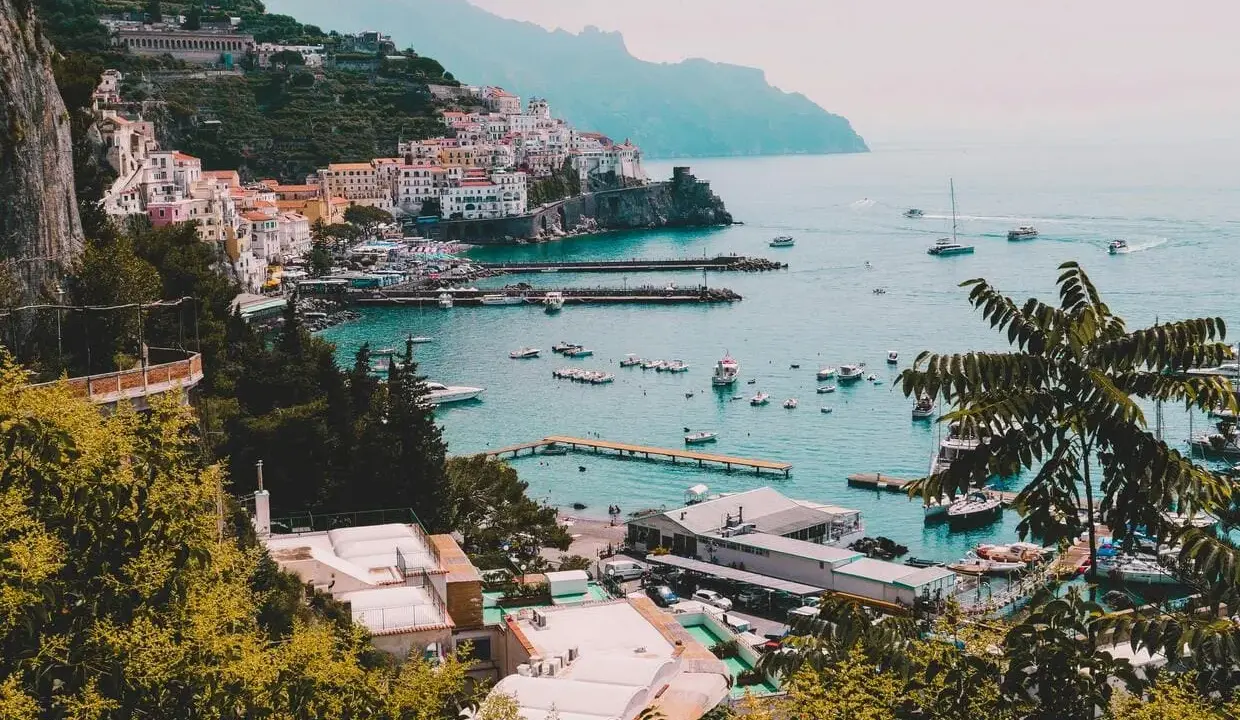 Vista panoramica di un villaggio colorato sulla Costiera Amalfitana, con barche nel porto e montagne sullo sfondo. Un invito a scoprire come visitare la Costiera Amalfitana.