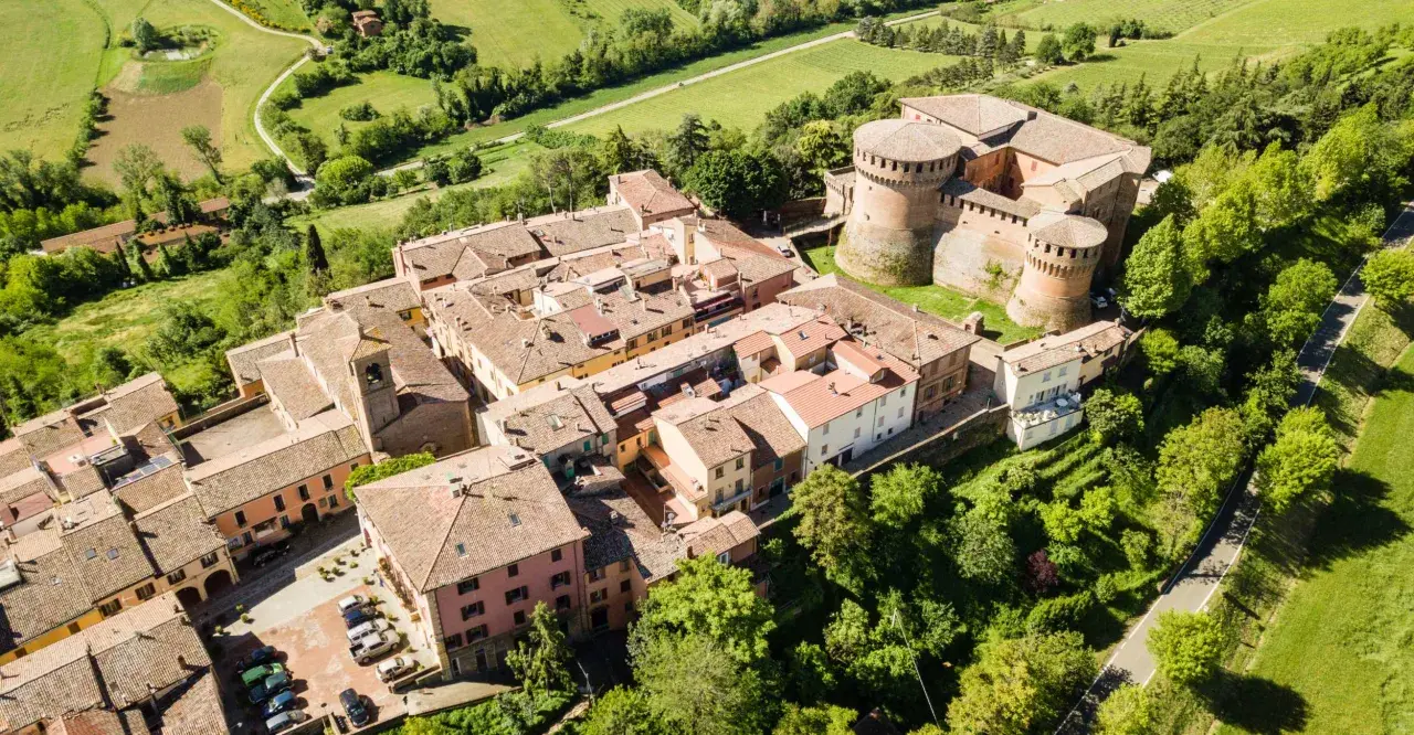 Aerial view of a medieval Italian village with a castle, terracotta roofs, and surrounding green hills.