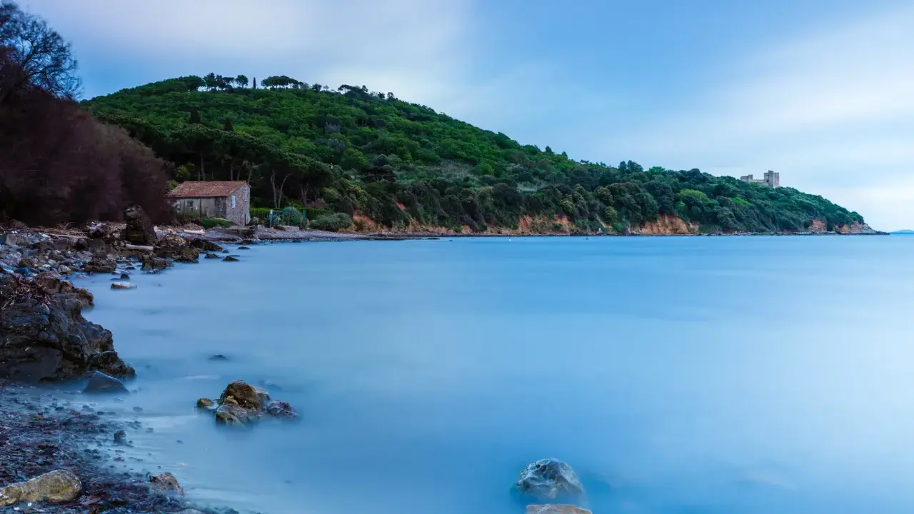 Una casa di pietra sulla spiaggia di Bengodi, con il mare calmo e una collina verdeggiante sullo sfondo.