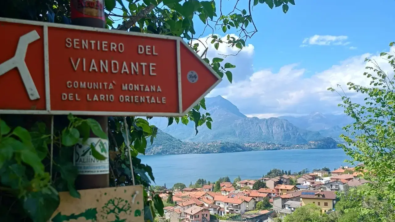 Segnale arancione indica "Sentiero del Viandante" con vista su un lago e montagne.