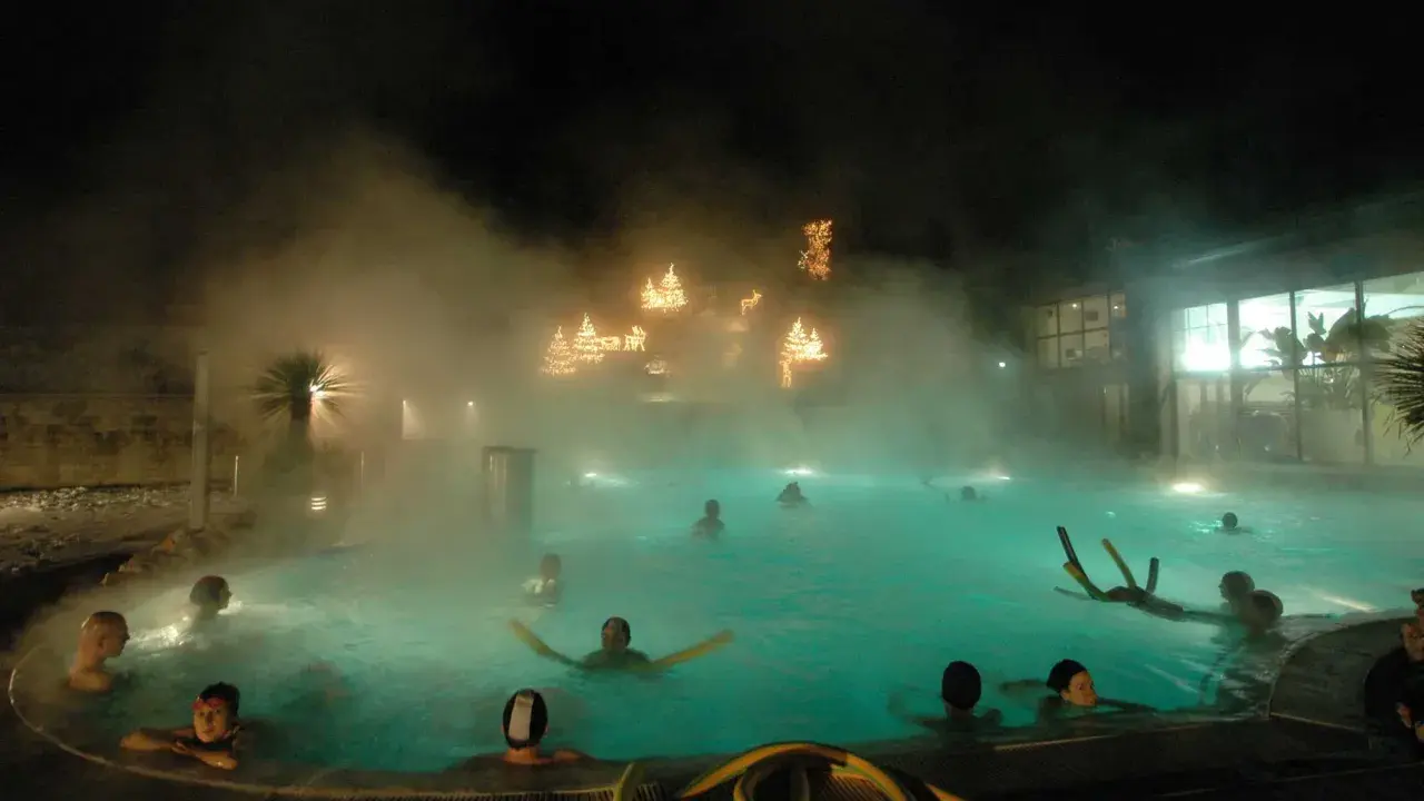 Persone si rilassano in una piscina termale notturna, con luci natalizie che brillano tra la nebbia, un'atmosfera magica nei bagni di Romagna.