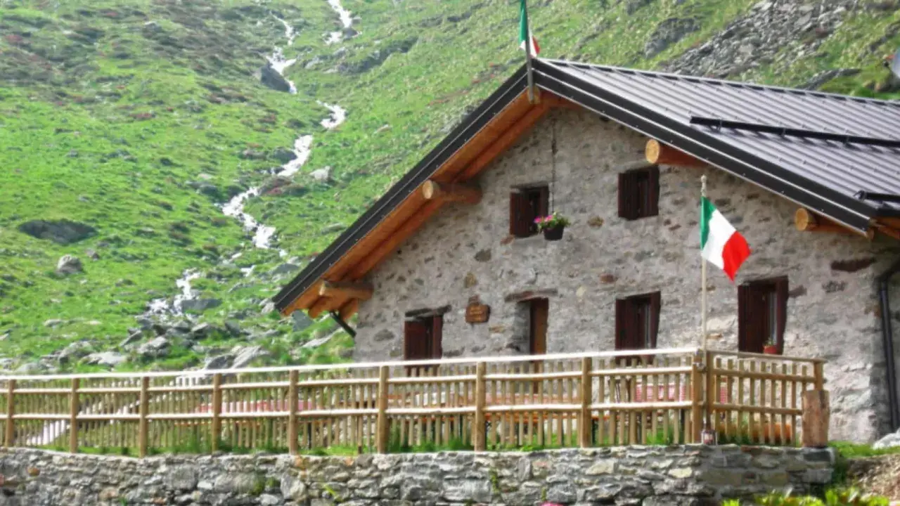 Rifugio in pietra con bandiera italiana, circondato da verdi pendii e un ruscello. Un'oasi accogliente tra i rifugi Val Camonica facili da raggiungere.
