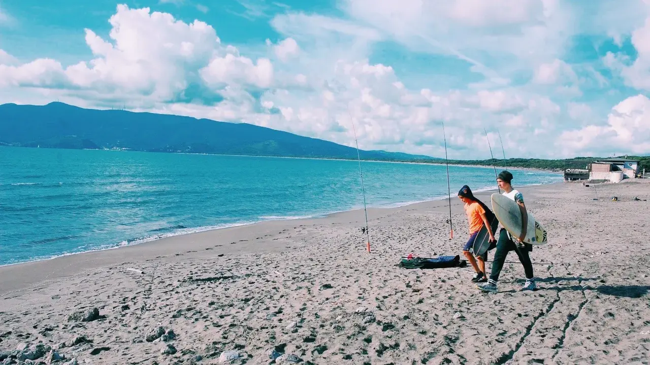 Due ragazzi passeggiano sulla spiaggia della Maremma Toscana, uno con una tavola da surf, mentre il mare azzurro lambisce la riva.