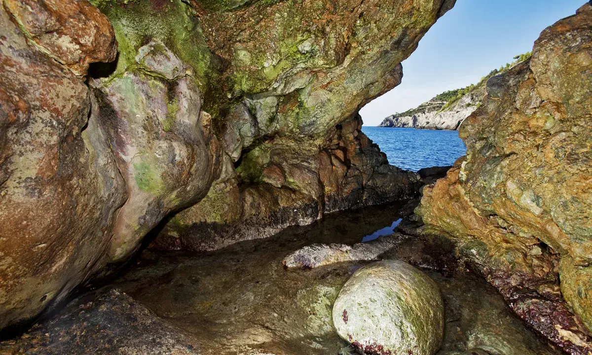 L'entrata di una grotta marina, la cala del bove, con rocce muschiose e un piccolo specchio d'acqua che riflette il blu del mare e il cielo.