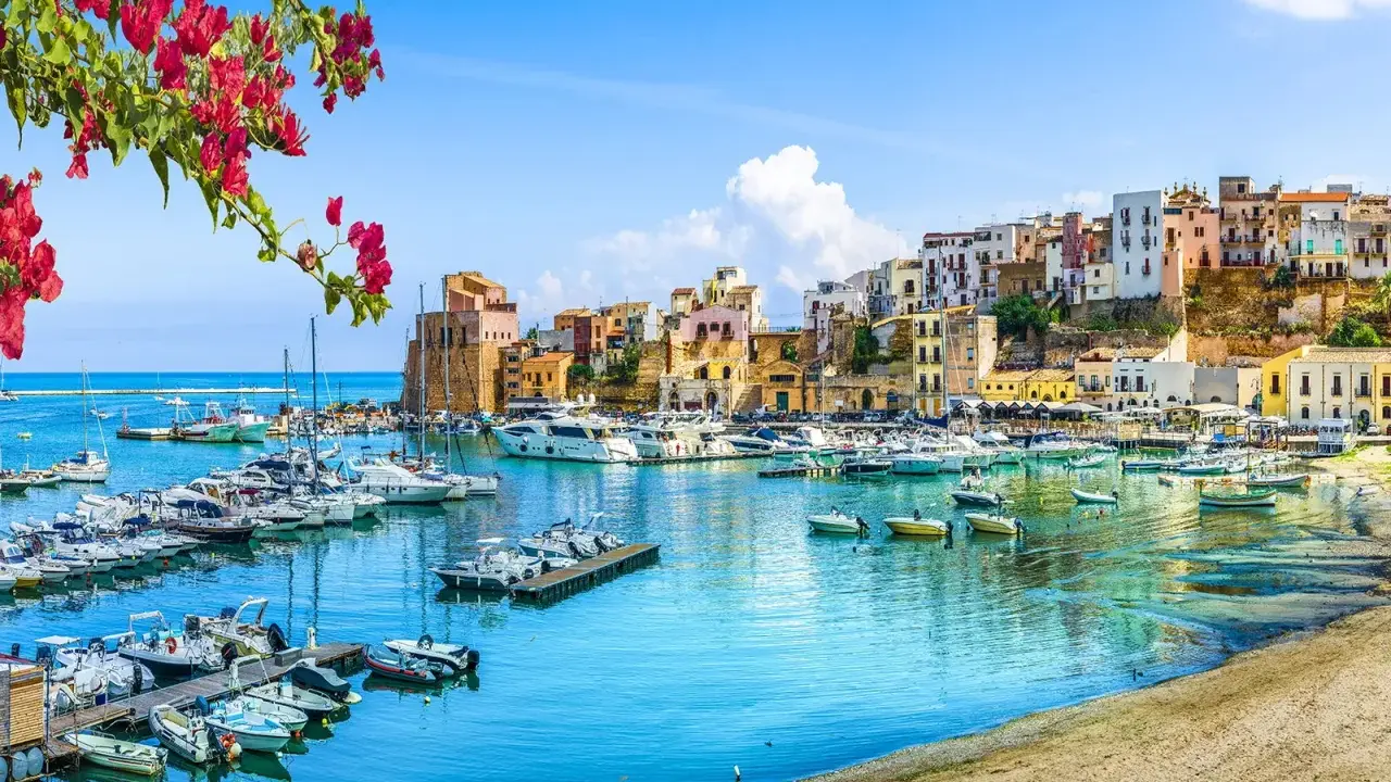 Un pittoresco porto con barche ormeggiate, case colorate su una collina e fiori rossi in primo piano, un'immagine che evoca la bellezza di Sardegna e Sicilia.