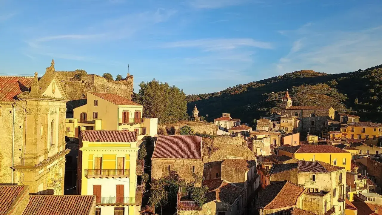 Panoramica di Castiglione di Sicilia al tramonto, con case dai tetti rossi e un castello in rovina su una collina.