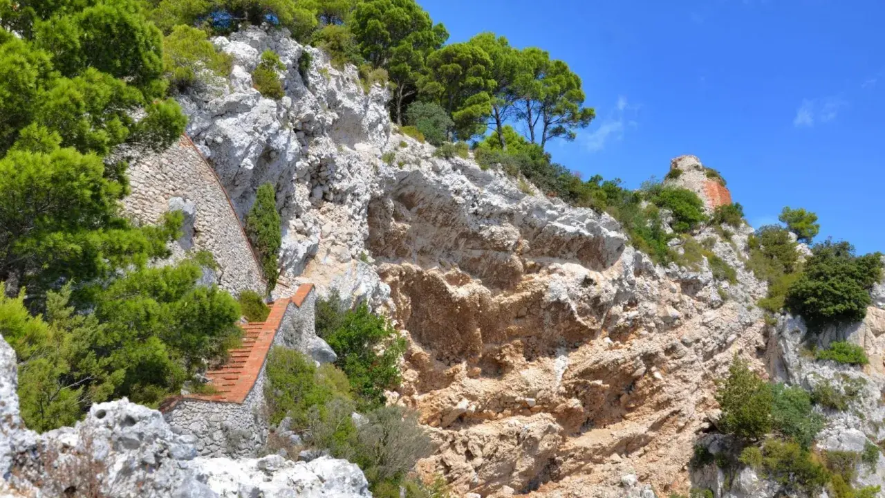 Scogliera rocciosa con una scalinata in mattoni che sale verso la cima, dove si intravede un rudere. Si immagina il **salto di Tiberio** da questo luogo suggestivo.