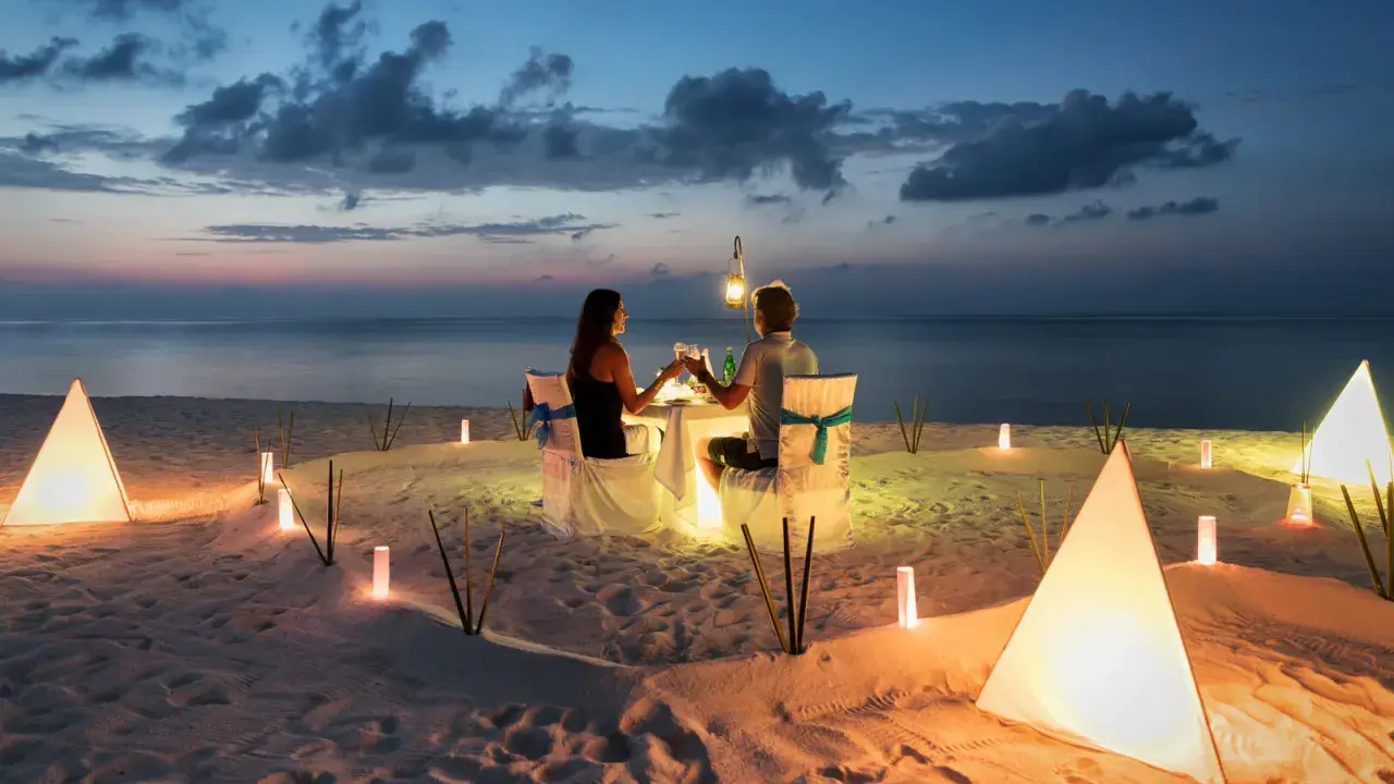 Una coppia al mare di sera, seduta a tavola per una cena romantica illuminata da lanterne e luci soffuse sulla sabbia.