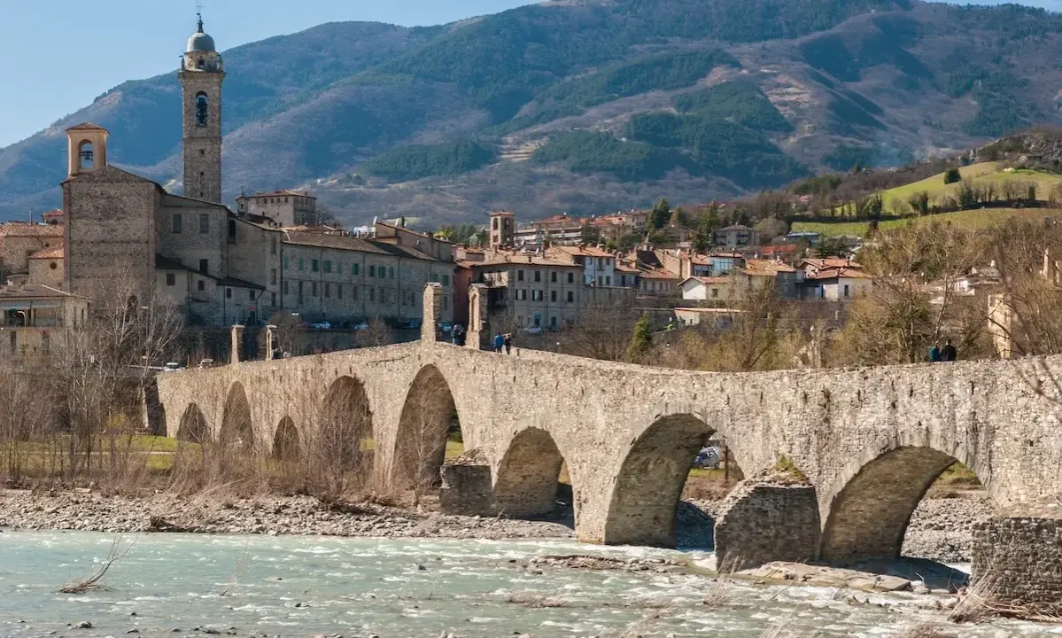 Ponte medievale in pietra attraversa un fiume, con un borgo storico e colline sullo sfondo. Un gioiello tra i borghi medievali dell'Emilia Romagna.