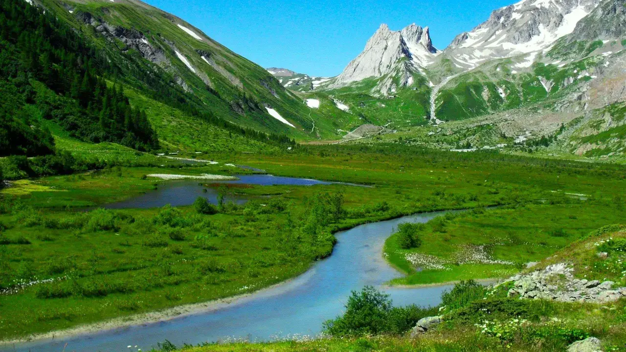 Val Ferret, un paradiso per passeggiate facili. Un fiume serpeggia tra prati verdi e montagne innevate sotto un cielo azzurro.