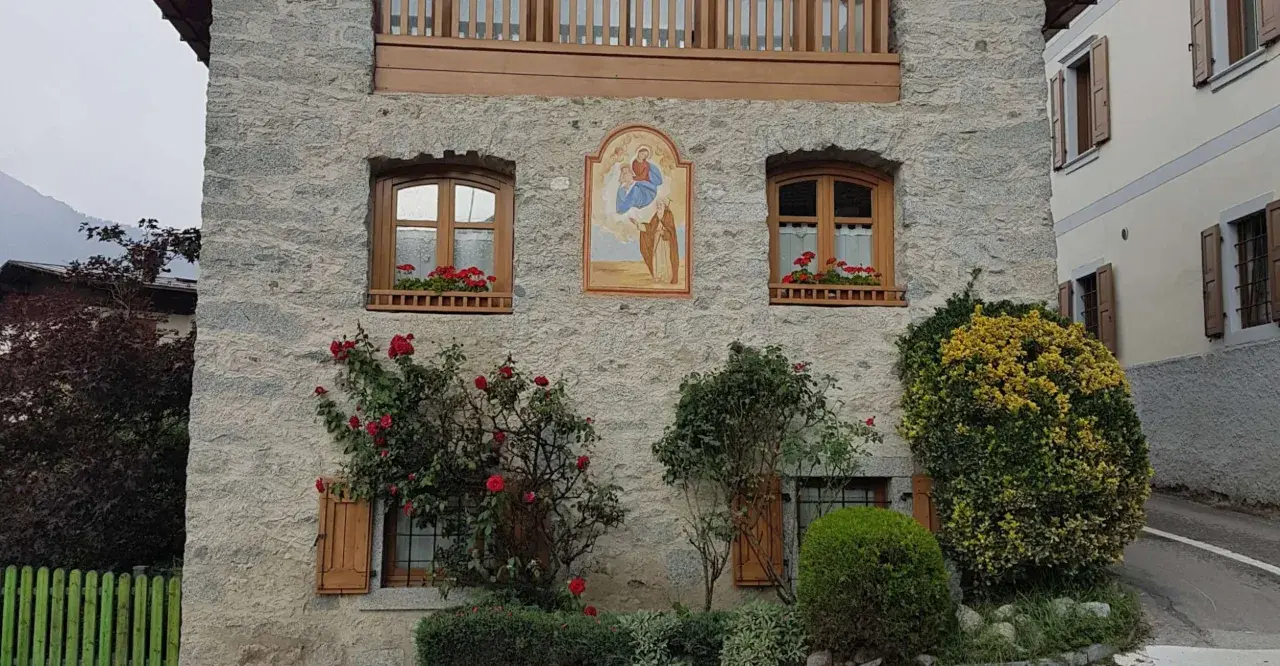 Un affresco sacro adorna un muro di pietra di un borgo trentino alto adige, con rose rosse che sbocciano rigogliose.