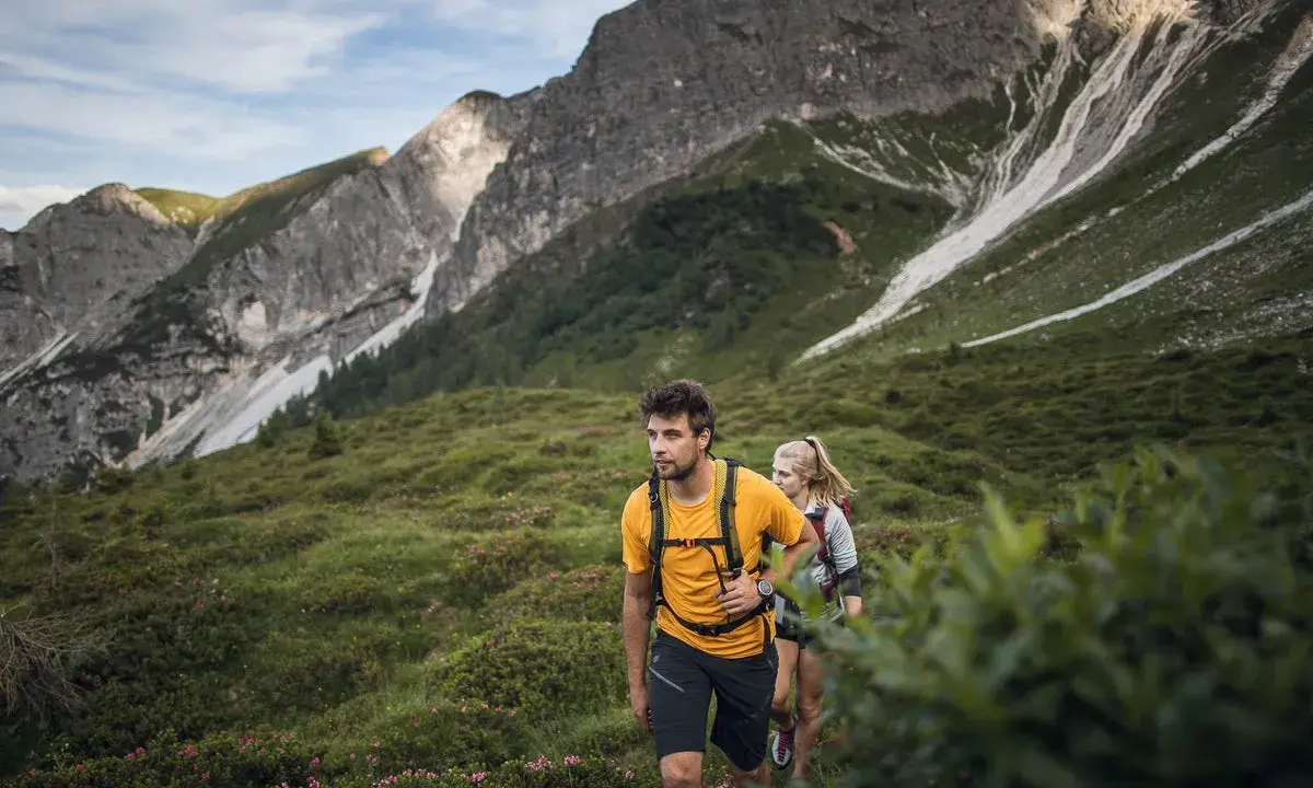 Coppia in escursioni alto adige, camminano su un sentiero verdeggiante con montagne imponenti sullo sfondo.