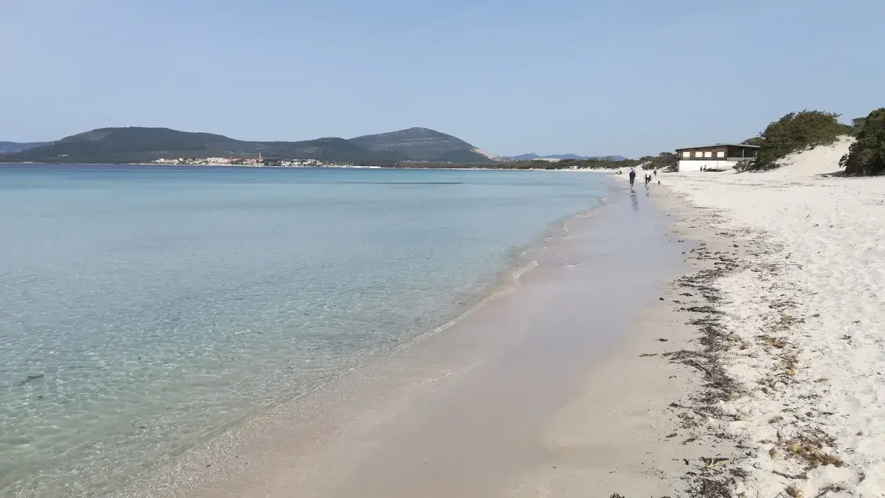 Spiaggia della Fertilia con acque cristalline, sabbia bianca e un edificio in lontananza. Due persone passeggiano lungo la riva.