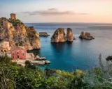 Un borgo con torri antiche e una casa rossa si affaccia su un mare bello Italia, con faraglioni che emergono dalle acque cristalline.