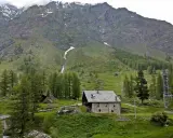 Un casolare in pietra nel **Val di Rhemes**, circondato da verdi pendii e imponenti montagne innevate.
