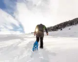 Uomo con ciaspole cammina su sentiero innevato, ideale per chi cerca dove ciaspolare in Piemonte. Sole splendente tra le nuvole.