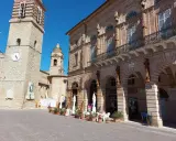 Piazza a San Ginesio, Marche, con il campanile in pietra e l'edificio in mattoni rossi con portici.