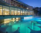 Piscina termale illuminata di notte, con vapori che salgono. Un edificio moderno con ampie vetrate si affaccia sull'acqua turchese, evocando un'atmosfera di relax e benessere, come in antiche terme preistoriche a Padova.