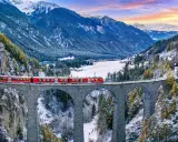Il trenino del Bernina attraversa un viadotto in pietra in un paesaggio alpino innevato, con montagne maestose e un cielo colorato al tramonto.