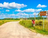 Segnaletica per la Via Francigena, un antico cammino. La strada sterrata conduce verso un borgo collinare sotto un cielo azzurro con nuvole bianche.