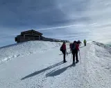 Gruppo di escursionisti con ciaspole su un sentiero innevato, con un rifugio in legno in cima a una collina. Posti belli in montagna Piemonte inverno.