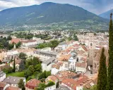 Vista aerea del centro storico di Merano, con montagne sullo sfondo e un campanile che svetta tra i tetti rossi.