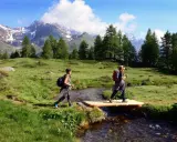 Due escursioniste attraversano un ruscello su un ponte di legno, godendosi un'attività in montagna tra prati verdi e cime innevate.