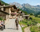Un escursionista cammina tra i pittoreschi paesi della Val di Non, con case in pietra e fiori colorati, mentre sullo sfondo si stagliano maestose montagne.