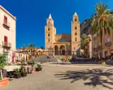 Piazza soleggiata con la cattedrale normanna sullo sfondo, palme e palazzi storici. Un luogo incantevole tra i borghi da visitare in Sicilia.