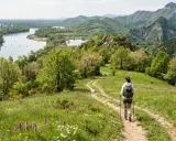 Donna con zaino e bastoncini da trekking su sentiero fiorito, godendosi le escursioni in Emilia Romagna con vista su fiume e montagne.