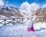 Bambina felice lancia neve in aria, con montagne innevate sullo sfondo. Perfetto per scoprire dove andare per la neve in Abruzzo.