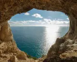 Vista dal mare da una grotta, con un sentiero del pellegrino che si intravede tra le rocce. Il sole brilla sull'acqua.