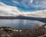 Vista panoramica del lago di Castel Gandolfo, Italia, con colline boscose e un cielo nuvoloso.