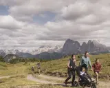 Famiglia in escursione sull'Alpe di Siusi, con bambini e passeggino, sullo sfondo le maestose vette dolomitiche.