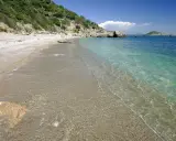 Spiaggia di ciottoli e sabbia con acqua cristallina, vista verso il mare aperto e un'isola in lontananza, vicino al porto di Ercole.