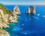 Scogliere verdi e rocciose si affacciano sul blu intenso del golfo di Napoli, punteggiato da barche e yacht. Faraglioni iconici emergono dal mare.