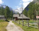 Sentiero di montagna con baite in pietra e tettoie, ideale per cogne escursioni facili. Montagne innevate sullo sfondo.