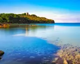 Torre medievale su una collina verdeggiante affacciata sul limpido **golfo di Follonica**. Acque calme e trasparenti riflettono il cielo azzurro.