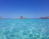 Acque cristalline del mare nord Italia con torre antica su isolotto e barca a vela arancione.