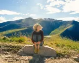 Ragazza seduta su una roccia, con i capelli mossi dal vento, ammira il panorama montano. Un momento di pace mentre camminare nella natura.